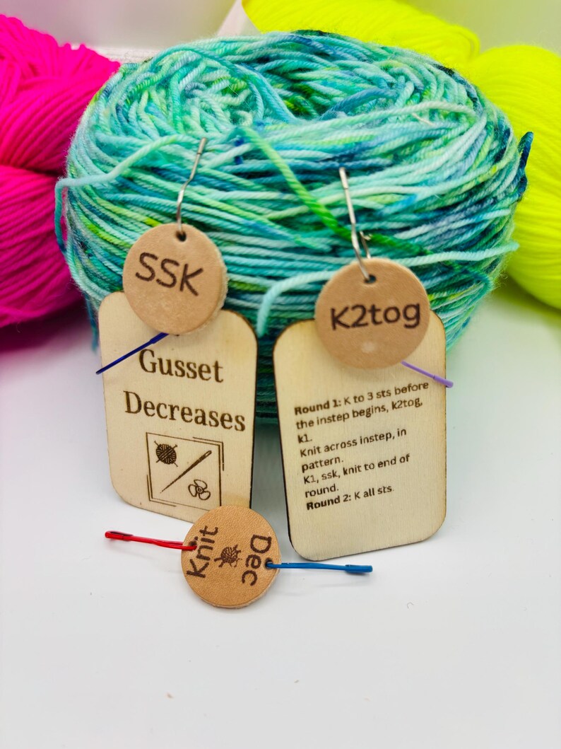 May include: Wooden knitting stitch markers with text including "SSK", "K2tog", "Gusset Decreases", "Knit Dec", and knitting instructions. The markers are attached to a blue and green yarn ball, with pink and yellow yarn in the background.