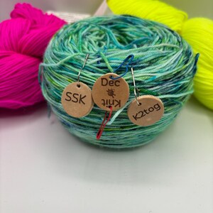 May include: A ball of variegated green and blue yarn with three small wooden tags attached. The tags read "SSK", "Dec Knit", and "K2tog". Bright pink and yellow yarn are visible in the background, suggesting a knitting or crochet theme.