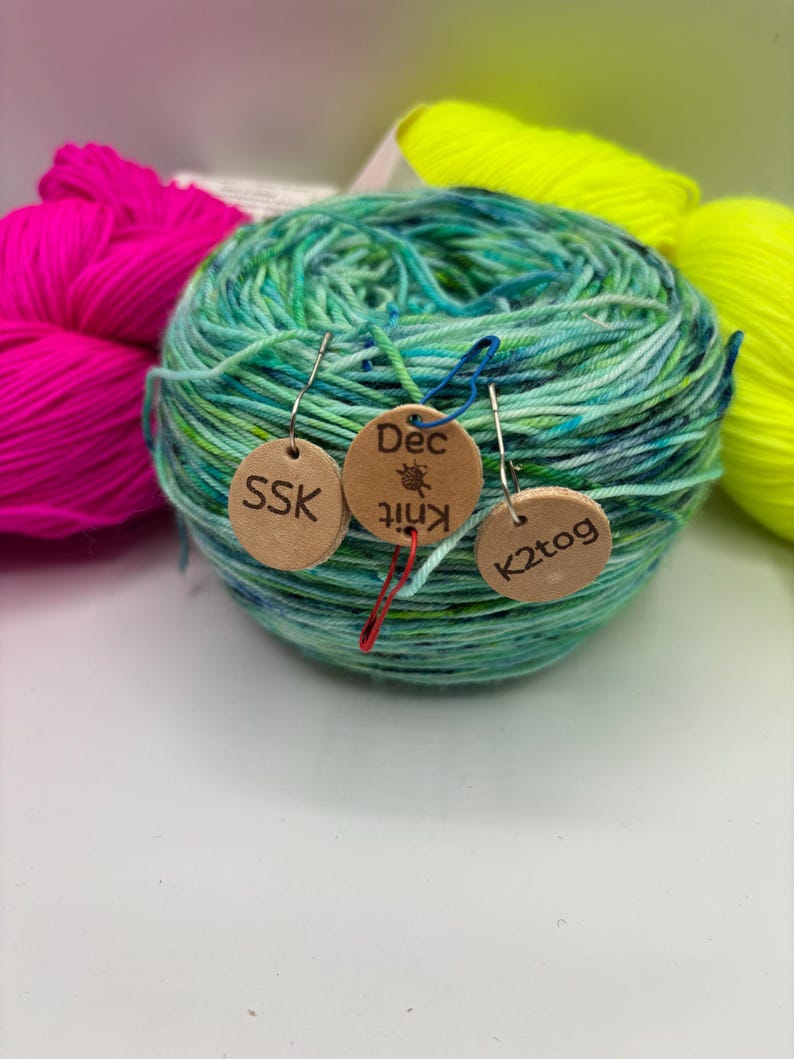 May include: A ball of variegated green and blue yarn with three small wooden tags attached. The tags read "SSK", "Dec Knit", and "K2tog". Bright pink and yellow yarn are visible in the background, suggesting a knitting or crochet theme.