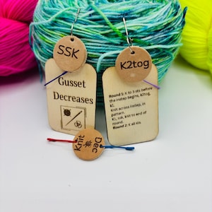May include: Wooden knitting stitch markers with text, including "SSK", "K2tog", and "Knit Dec". The markers are attached to small tags with instructions for "Gusset Decreases". Colourful yarn in shades of blue, green, pink, and yellow is in the background.