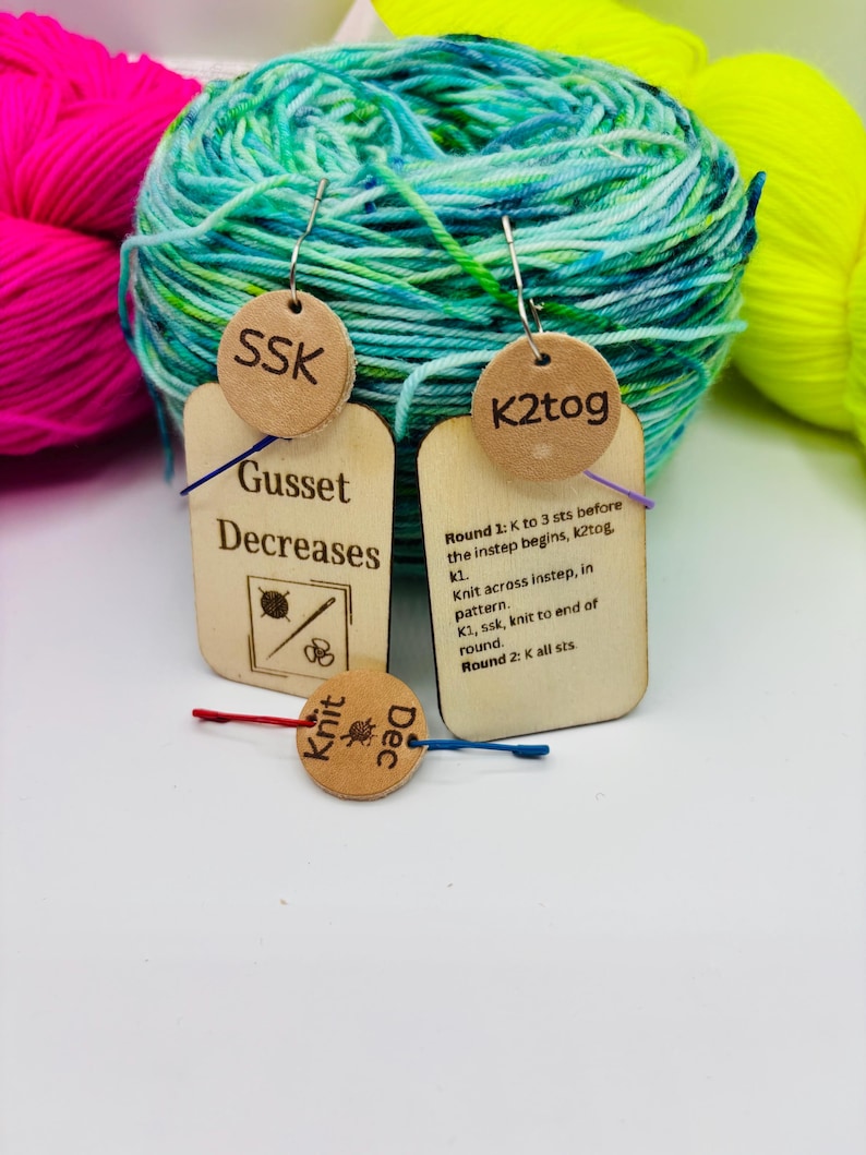 May include: Wooden knitting stitch markers with text, including "SSK", "K2tog", and "Knit Dec". The markers are attached to small tags with instructions for "Gusset Decreases". Colorful yarn in shades of blue, green, pink, and yellow is in the background.