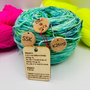May include: A ball of teal and green yarn with three wooden tags attached. The tags have the abbreviations "SSK" and "K2tog", and the word "Dec Knit". A larger tag has knitting instructions.