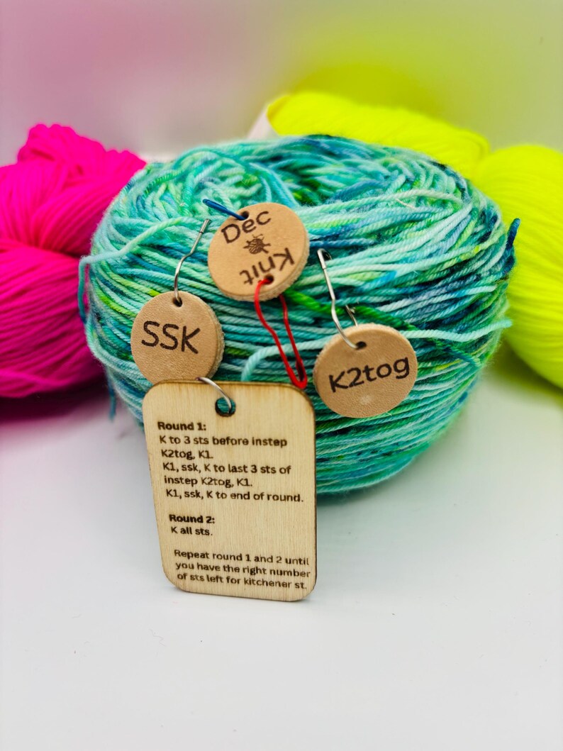 May include: A ball of teal and green yarn with three wooden tags attached. The tags have the abbreviations "SSK" and "K2tog", and the word "Dec Knit". A larger tag has knitting instructions.