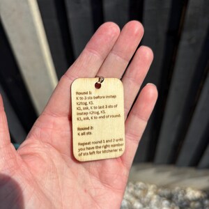 May include: A small, rectangular wooden tag with a metal loop at the top. The tag features printed knitting instructions. The text includes terms like "K2tog" and "ssk". The tag is held in a person's hand.