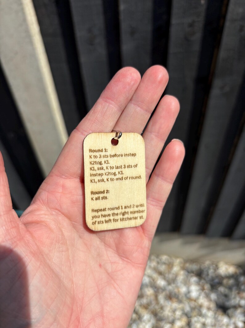 May include: A small, rectangular wooden tag with a metal loop at the top. The tag features printed knitting instructions. The text includes terms like "K2tog" and "ssk". The tag is held in a person's hand.