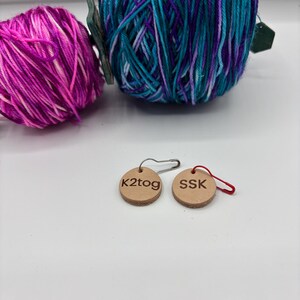 May include: Two balls of yarn in shades of pink, purple, and teal. Two small, round, wooden tags with the text "k2tog" and "SSK" are also visible. The tags are attached to safety pins and red loops.
