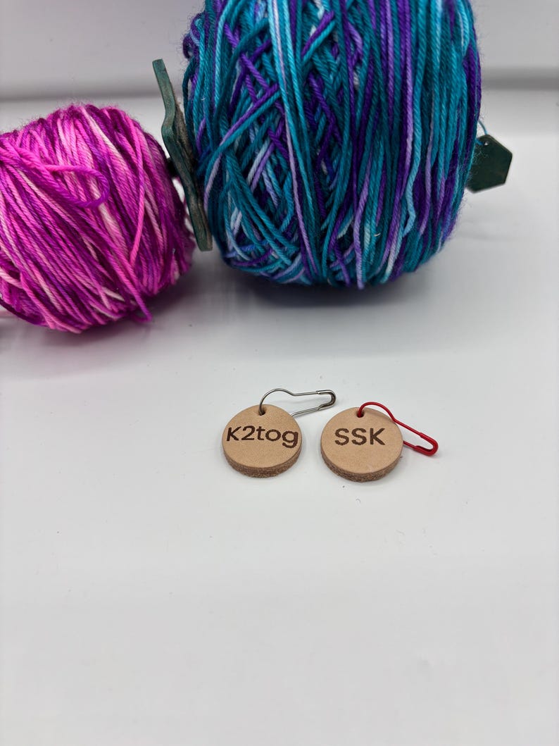 May include: Two balls of yarn in shades of pink, purple, and teal. Two small, round, wooden tags with the text "k2tog" and "SSK" are also visible. The tags are attached to safety pins and red loops.