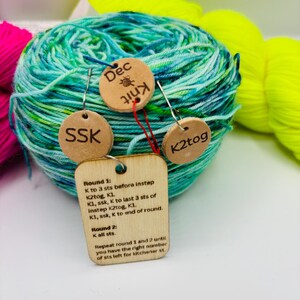 May include: A ball of teal and green yarn with wooden tags attached. The tags have the words "SSK", "Dec Knit", and "K2tog" engraved on them. A larger tag displays knitting instructions. Other yarn skeins in pink and yellow are in the background.