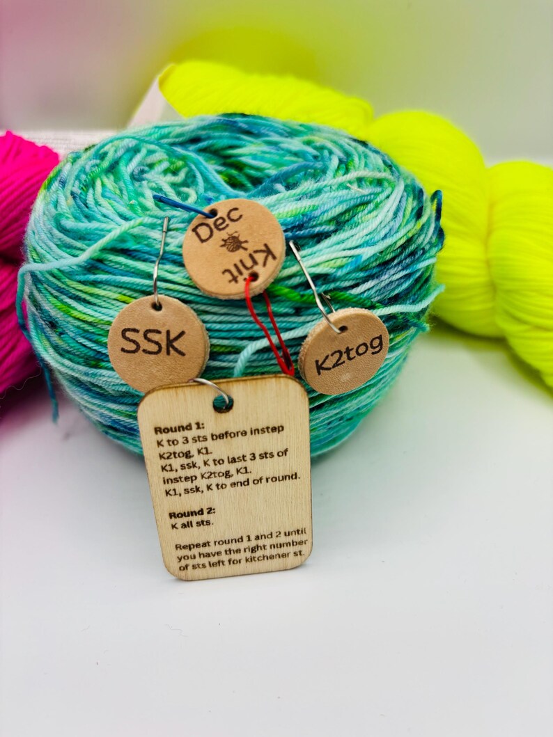 May include: A ball of teal and green yarn with wooden tags attached. The tags have the words "SSK", "Dec Knit", and "K2tog" engraved on them. A larger tag displays knitting instructions. Other yarn skeins in pink and yellow are in the background.
