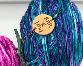 May include: A close-up of two balls of yarn. One ball is a vibrant mix of teal, purple, and white yarn, with a wooden tag that reads "Knit Purl". A green needle and a red stitch marker are attached to the yarn. The other ball is pink and purple.
