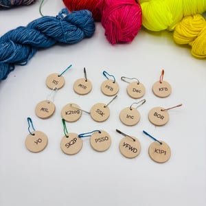 May include: Wooden stitch markers with abbreviations like RS, M1R, and K2tog, attached to safety pins. The markers are arranged in front of colourful yarn skeins in blue, red, pink, and yellow. Ideal for knitting projects.