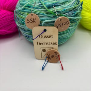May include: A ball of teal and green yarn with small wooden tags attached. The tags have the text "SSK", "K2tog", "Gusset Decreases", and "Knit Dec". Other balls of yarn in pink and yellow are in the background.