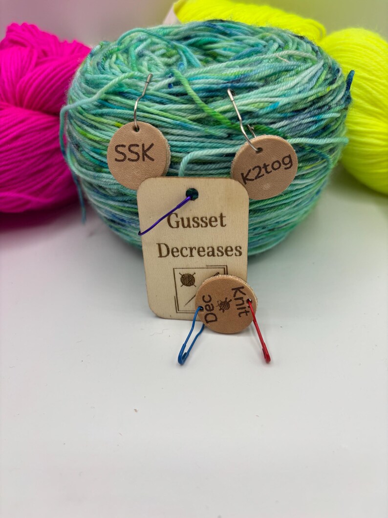 May include: A ball of teal and green yarn with small wooden tags attached. The tags have the text "SSK", "K2tog", "Gusset Decreases", and "Knit Dec". Other balls of yarn in pink and yellow are in the background.