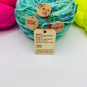 May include: A ball of teal and green variegated yarn with wooden tags attached. The tags read "Dec Knit", "SSK", and "2tog". A larger tag displays knitting instructions. Bright pink and yellow yarn are in the background.