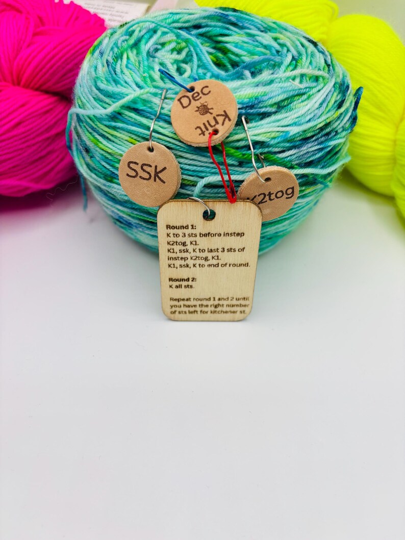 May include: A ball of teal and green variegated yarn with wooden tags attached. The tags read "Dec Knit", "SSK", and "2tog". A larger tag displays knitting instructions. Bright pink and yellow yarn are in the background.