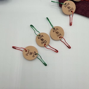 May include: Circular, light brown leather knitting stitch markers with the words "Knit" and "Dec" or "Purl" printed on them. Each marker has a safety pin attached, with green and red pins. A ball of blue yarn is visible in the background.