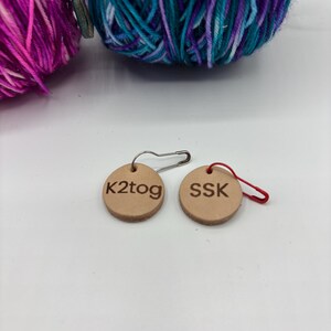 May include: Two small, round, tan leather stitch markers with the text "K2tog" and "SSK". One marker has a silver safety pin, the other a red loop. Two balls of yarn in shades of purple, teal, and blue are in the background.