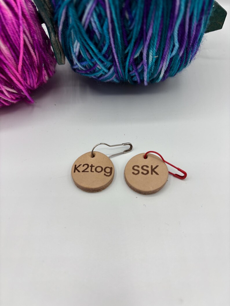 May include: Two small, round, tan leather stitch markers with the text "K2tog" and "SSK". One marker has a silver safety pin, the other a red loop. Two balls of yarn in shades of purple, teal, and blue are in the background.