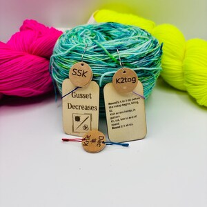May include: A collection of colorful yarn skeins and knitting stitch markers. The yarn is in shades of pink, green, and yellow. The stitch markers are small, round, and made of wood, with text such as "SSK" and "K2tog".