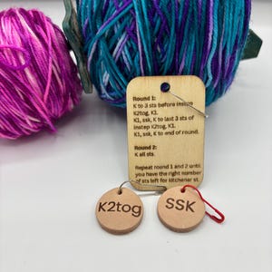 May include: Image of knitting supplies featuring two balls of yarn in purple, teal, and pink. A wooden tag displays knitting instructions, and two small, round leather tags show the abbreviations "K2tog" and "SSK".