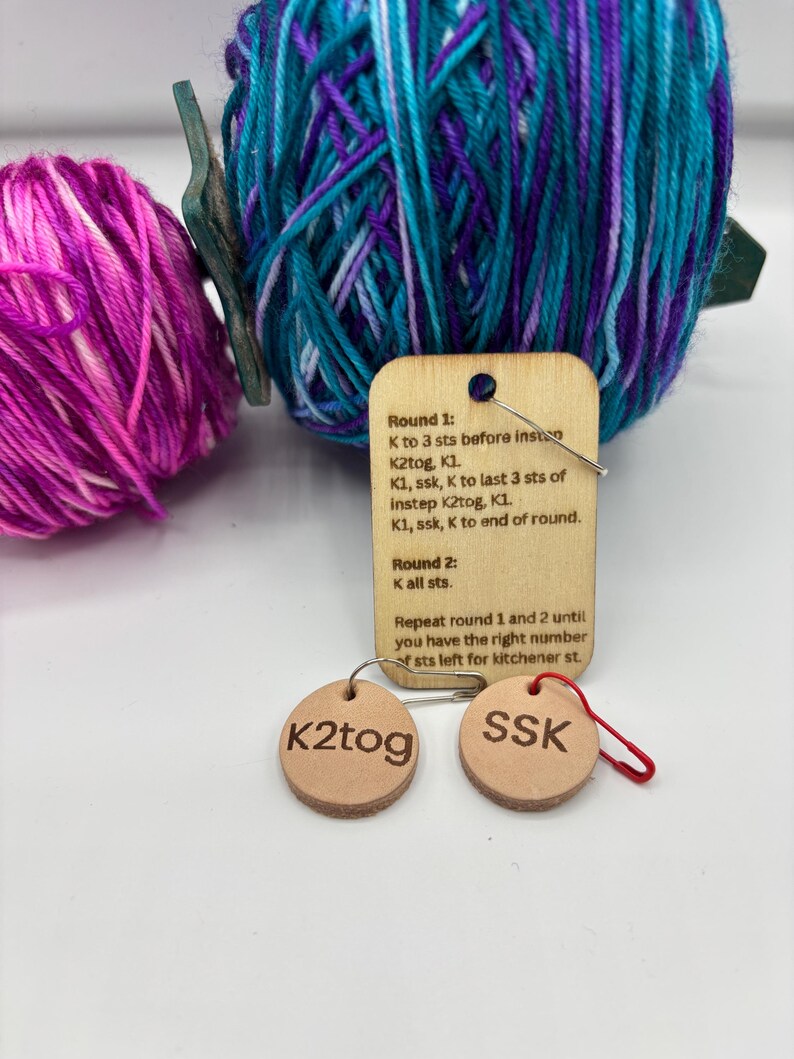 May include: Image of knitting supplies featuring two balls of yarn in purple, teal, and pink. A wooden tag displays knitting instructions, and two small, round leather tags show the abbreviations "K2tog" and "SSK".