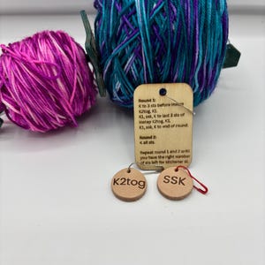 May include: A close-up of knitting supplies, including two skeins of yarn in shades of purple, pink, and teal. Two small, round leather tags with the text "K2tog" and "SSK" are attached with a safety pin. A wooden tag with knitting instructions is also present.