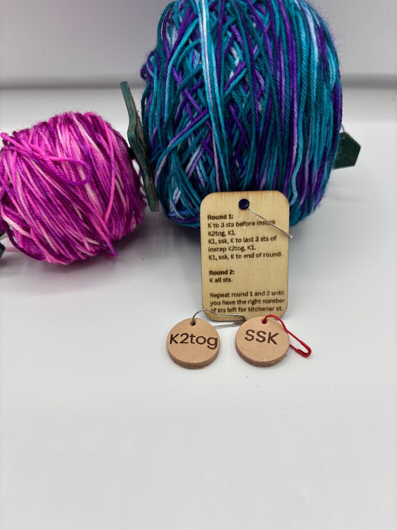 May include: A close-up of knitting supplies, including two skeins of yarn in shades of purple, pink, and teal. Two small, round leather tags with the text "K2tog" and "SSK" are attached with a safety pin. A wooden tag with knitting instructions is also present.