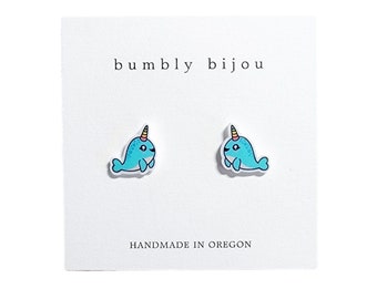 Peut inclure: Une paire de boucles d'oreilles clous en forme de narval bleu avec une corne jaune et rose. Les boucles d'oreilles sont sur une carte blanche avec le texte "bumbly bijou" et "HANDMADE IN OREGON".