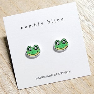 May include: A pair of green frog earrings with white outlines and red cheeks. The earrings are on a white card with the text "bumbly bijou" and "HANDMADE IN OREGON".
