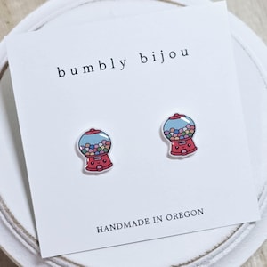 May include: A pair of red gumball machine earrings with a smiling face and colorful gumballs inside. The earrings are on a white card with the text "bumbly bijou" and "HANDMADE IN OREGON".