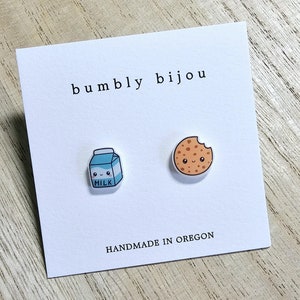May include: A white card with the text "bumby bijou" and "HANDMADE IN OREGON" printed on it. There are two small cartoon earrings on the card: a blue milk carton with a smiling face and a brown cookie with a smiling face.