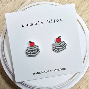 May include: A pair of earrings featuring a stack of books with a red apple on top. The earrings are handmade in Oregon and are sold by the shop "bumbly bijou".