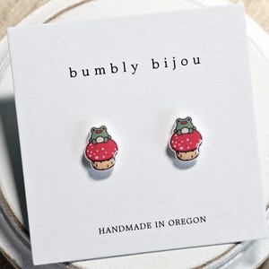 May include: A pair of stud earrings featuring a red mushroom design with white polka dots and a small green frog on top. The earrings are displayed on a white card with the text "bumbly bijou" and "HANDMADE IN OREGON".