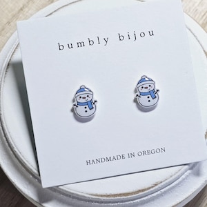 May include: A pair of snowman stud earrings with blue hats and scarves. The earrings are on a white card with the text "bumbly bijou" and "HANDMADE IN OREGON".