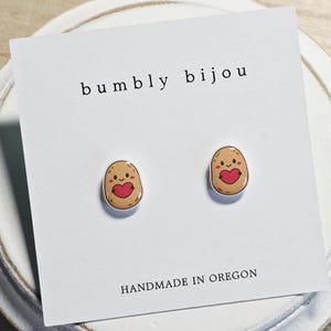 May include: A pair of potato-shaped stud earrings with a red heart design. The earrings are on a white card with the text "bumbly bijou" and "HANDMADE IN OREGON". The earrings are set against a white background.