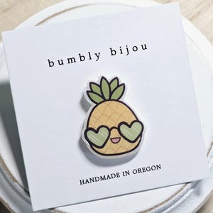 May include: A yellow pineapple with a green top and heart-shaped sunglasses, with the text "bumbly bijou" and "HANDMADE IN OREGON" on a white card.