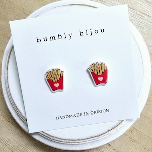 May include: A pair of white stud earrings with a red and yellow french fry design. The earrings have a small pink heart on the front. The earrings are on a white card with the text "bumbly bijou" and "HANDMADE IN OREGON".