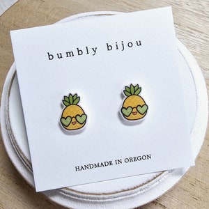 May include: A pair of yellow pineapple earrings with green leaves and heart-shaped sunglasses. The earrings are on a white card with the text "bumbly bijou" and "HANDMADE IN OREGON".