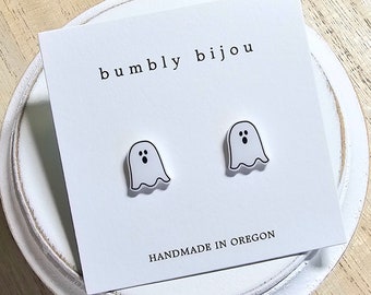 May include: A pair of white ghost-shaped earrings with black eyes on a white card with the text "bumbly bijou" and "HANDMADE IN OREGON".