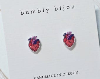 May include: A pair of stud earrings featuring anatomical heart illustrations in shades of pink, red, and purple. The earrings are attached to a white display card printed with the text bumbly bijou at the top and HANDMADE IN OREGON at the bottom.