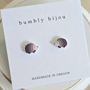May include: A pair of stud earrings featuring a small, illustrated hedgehog design in shades of brown and cream. The earrings are attached to a white display card printed with the text bumbly bijou at the top and HANDMADE IN OREGON at the bottom.