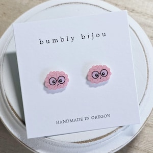 May include: A pair of pink brain-shaped stud earrings with smiling faces and glasses. The earrings are displayed on a white card with the text "bumbly bijou" and "HANDMADE IN OREGON".