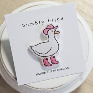 May include: A white duck pin wearing a pink cowboy hat and pink cowboy boots. The pin is on a white card with the text "bumbly bijou" and "HANDMADE IN OREGON". The duck has a yellow beak and black eyes.