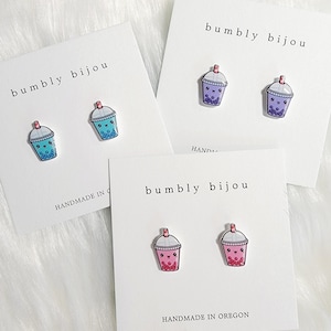 May include: Three pairs of bubble tea earrings. The first pair is blue and white, the second pair is purple and white, and the third pair is pink and white. Each pair has a cute cartoon face on the bubble tea cup. The cards say "bumbly bijou" and "handmade in Oregon".