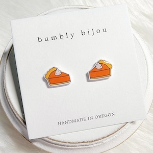 May include: A pair of pumpkin pie slice earrings with a white swirl of whipped cream on top. The earrings are on a white card with the text "bumbly bijou" and "HANDMADE IN OREGON".