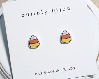 May include: A pair of stud earrings shaped like candy corn with white, orange, and yellow horizontal stripes. The earrings are attached to a white display card featuring the text bumbly bijou at the top and HANDMADE IN OREGON at the bottom.