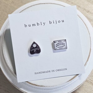May include: A pair of silver earrings with a black and white Ouija board design and a black and white "Goodbye" design. The earrings are on a white card with the text "bumby bijou" and "Handmade in Oregon".