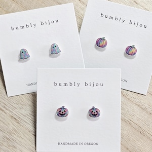 May include: Three sets of stud earrings on white display cards labeled bumbly bijou. One set features iridescent ghost shapes, another shows iridescent pumpkins, and the third displays iridescent jack-o'-lanterns with carved faces.