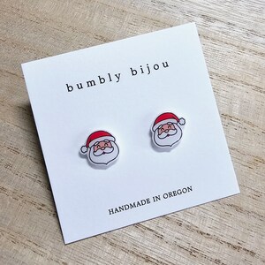 Peut inclure: Une paire de boucles d'oreilles blanches en forme de P&egrave;re No&euml;l avec des chapeaux rouges. Les boucles d'oreilles sont sur une carte blanche avec le texte "bumbly bijou" et "HANDMADE IN OREGON".
