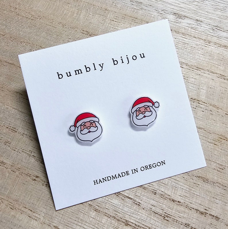Peut inclure: Une paire de boucles d'oreilles blanches en forme de P&egrave;re No&euml;l avec des chapeaux rouges. Les boucles d'oreilles sont sur une carte blanche avec le texte "bumbly bijou" et "HANDMADE IN OREGON".